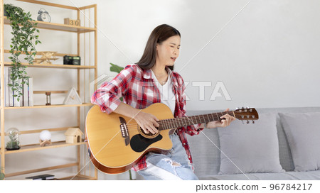 Asian female artist playing the guitar and singing happily in the living room, Relaxation with music therapy, Spending free time with music, Joy of playing music, Acoustic guitar. 96784217