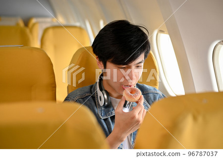 Relaxed Asian male passenger is eating a doughnut during the flight. Airplane concept 96787037