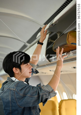Handsome young Asian male passenger putting his baggage into an overhead locker. 96787038