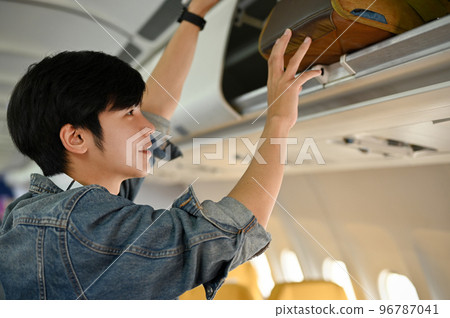 Handsome Asian male passenger putting his carry-on baggage into an overhead locker. Handsome Asian male passenger putting his carry-on baggage into an overhead locker. 96787041