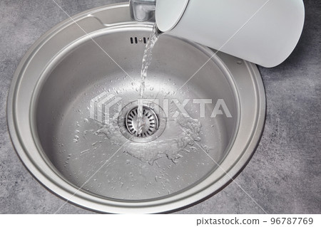 Hot water pours from a white electric kettle into a metal kitchen sink. Debris cleaning and disinfection Hot water pours from a white electric kettle into a metal kitchen sink. Debris cleaning and disinfection 96787769