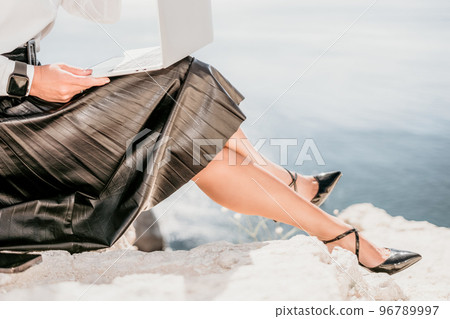 Digital nomad, Business woman working on laptop by the sea. Pretty lady typing on computer by the sea at sunset, makes a business transaction online from a distance. Freelance, remote work on vacation 96789997