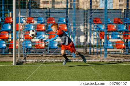 Junior goalkeeper catching ball while defending gate in football match Junior goalkeeper catching ball while defending gate in football match 96790407