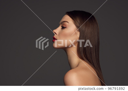 Profile view of female fashion model with naked shoulders and long hair looking at camera over dark grey background. Beautiful woman with trendy make-up, red lipstick and well-kept skin. 96791892