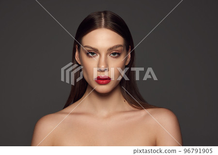 Portrait of female fashion model with naked shoulders and long hair looking at camera over dark grey background. Beautiful woman with trendy make-up, red lipstick and well-kept skin. Portrait of female fashion model with naked shoulders and long hair looking at camera over dark grey background. Beautiful woman with trendy make-up, red lipstick and well-kept skin. 96791905