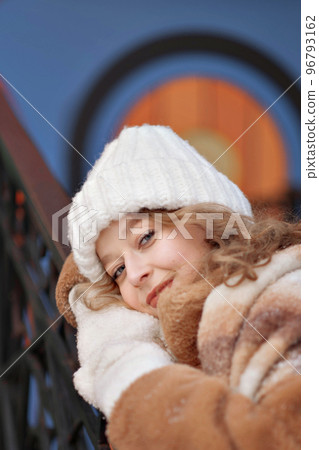 A white blonde girl in a knitted hat, a long scarf and a fur coat walks through the forest and smiles. Woman catches snow in white mittens. Winter is on the street. High quality photo. Christmas Eve 96793162