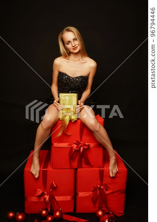 Woman gifts christmas. Beautiful sexy young blonde in a black dress sits on three big red gifts on a black background. Christmas photo, gifts 96794446