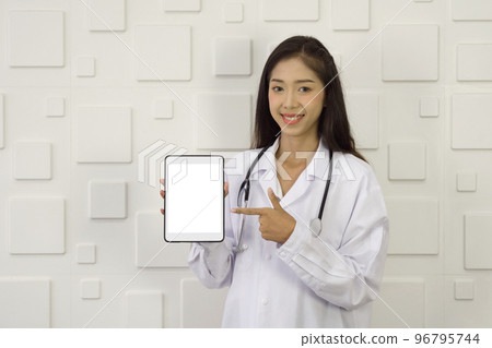 Young asian doctor in white gown and stethoscope pointing finger at white screen tablet computer, standing in front of white rectangle pattern ceramic wall. Young asian doctor in white gown and stethoscope pointing finger at white screen tablet computer, standing in front of white rectangle pattern ceramic wall. 96795744