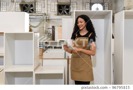 teenager in brown apron checking email order on tablet computer. Wooden cabinets of various sizes are placed in the background. Atmosphere in a wooden furniture factory. 96795815