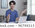 Asian man looking at camera while holding credit card and using laptop computer 96799313