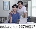 Asian gay couple smiling looking at camera while holding credit card and using laptop computer 96799317