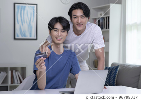Asian gay couple smiling looking at camera while holding credit card and using laptop computer Asian gay couple smiling looking at camera while holding credit card and using laptop computer 96799317