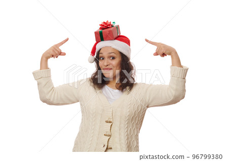 Overjoyed woman wearing Santa hat, looking at camera and pointing on Christmas present on her head, isolated over white background with advertising space. New Year, Xmas and winter holidays concept 96799380