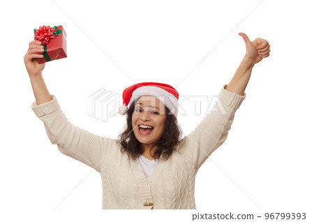 African American overjoyed pretty woman in Santa hat, holds a Christmas gift and shows thumb up, cutely smiling looking at camera, isolated on white background. New Year celebration and Xmas concept 96799393