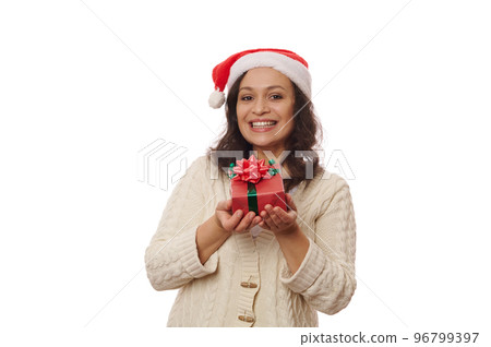 Multi-ethnic brunette, a happy middle-aged woman in Santa hat and beige sweater, showing a Christmas present in red wrapping paper and bow, cutely smiling, looking at camera, against white background Multi-ethnic brunette, a happy middle-aged woman in Santa hat and beige sweater, showing a Christmas present in red wrapping paper and bow, cutely smiling, looking at camera, against white background 96799397