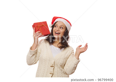 Delightful pretty woman in Santa hat, shaking a beautiful gift box, guessing what it contains, isolated over white background. Time to unpack Christmas and New Year's presents. Happy winter holidays 96799400