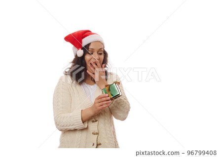Multi-ethnic beautiful woman in Santa hat received gift box with bow. She is happy and flattered by attention. Charming brunette smiling with present on white background. New year, Christmas concept. 96799408
