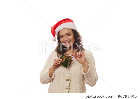 Attractive woman in Santa hat, looking at camera with seductive look, smiling with beautiful toothy smile, untying gold ribbon on Xmas New Years present, isolated on white background. Merry Christmas 96799409