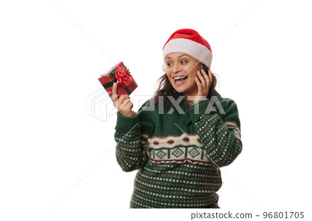 Multi-ethnic excited beautiful woman, wearing warm Xmas patterned sweater and Santa hat, smiling a cheerful toothy smile looking at her Christmas present, isolated over white background. Copy space 96801705