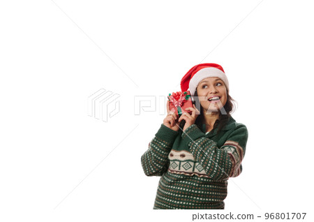 Latin American overjoyed pretty woman, in warm winter green sweater and Santa hat, shakes Christmas gift, guessing what is inside it, isolated on white background. Time to open presents. Boxing Day 96801707