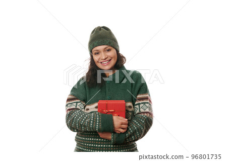 Merry Christmas. Portrait of a beautiful multi-ethnic woman in a cozy knitted green Xmas patterned sweater and wool green hat, holding gift box, smiling looking at camera, isolated on white background Merry Christmas. Portrait of a beautiful multi-ethnic woman in a cozy knitted green Xmas patterned sweater and wool green hat, holding gift box, smiling looking at camera, isolated on white background 96801735