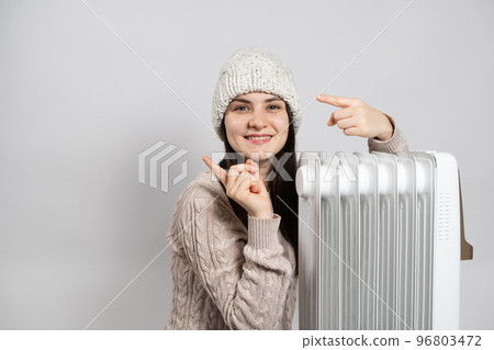 A woman basks near an electric oil heater and points her fingers to the side in place for text. Methods of heating an apartment or house. 96803472