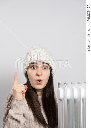 A woman in a hat getting warm near the oil heater and looks and points up with her finger. 96803475