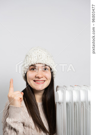 A woman in a hat getting warm near the oil heater and looks and points up with her finger. 96803477