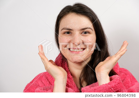 A young brunette woman in a pink bathrobe makes cleansing facial treatments - a mask or scrub to cleanse and nourish the skin. 96804398