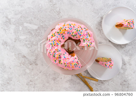 Flat lay. Sliced funfettti bundt cake with pink buttercream frosting on top and buttercream filling inside. 96804760