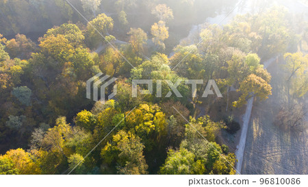 Aerial flying over trees with yellow leaves, meadow, dirt road with morning mist an autumn sunny morning in park. Bright sunlight, shining sunbeams rays, sun overexposure. Beautiful natural background 96810086