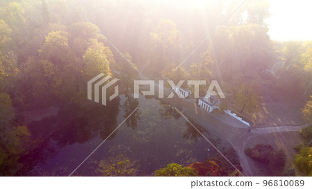 Aerial drone view over lake with swans, architectural columns and trees with yellow-green leaves in park on a bright sunny autumn morning. Sunbeams, red sun flare, sun overexposure. Natural background 96810089