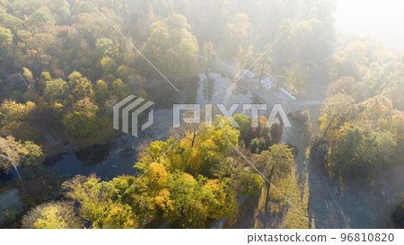 Aerial drone view over lake with swans, architectural columns and trees with yellow-green leaves in park on a bright sunny autumn morning. Sunbeams, red sun flare, sun overexposure. Natural background Aerial drone view over lake with swans, architectural columns and trees with yellow-green leaves in park on a bright sunny autumn morning. Sunbeams, red sun flare, sun overexposure. Natural background 96810820