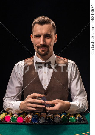 Portrait of a croupier is holding playing cards, gambling chips on table. Black background Portrait of a croupier is holding playing cards, gambling chips on table. Black background 96818674