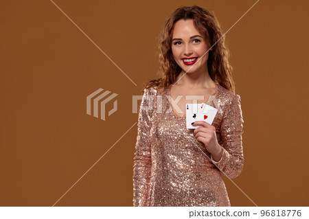 Pretty young brown-haired woman holding pair of aces in her hands wearing golden cocktail dress on brown background 96818776