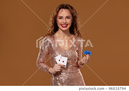Attractive brown-haired caucasian young woman in golden cocktail dress at casino holds pair of aces and chips in her hands Attractive brown-haired caucasian young woman in golden cocktail dress at casino holds pair of aces and chips in her hands 96818794