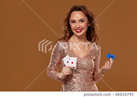 Attractive brown-haired caucasian young woman in golden cocktail dress at casino holds pair of aces and chips in her hands 96818795