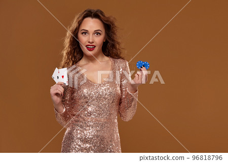 Attractive brown-haired caucasian young woman in golden cocktail dress at casino holds pair of aces and chips in her hands Attractive brown-haired caucasian young woman in golden cocktail dress at casino holds pair of aces and chips in her hands 96818796