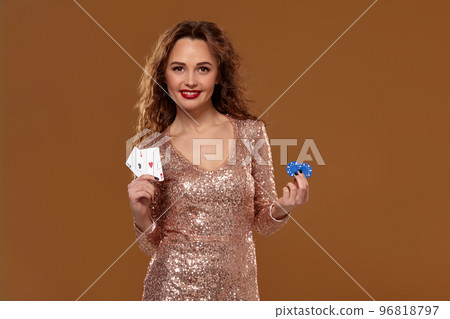 Attractive brown-haired caucasian young woman in golden cocktail dress at casino holds pair of aces and chips in her hands 96818797