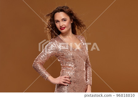 Portrait of young caucasian female in golden cocktail dress looking into the camera on brown background Portrait of young caucasian female in golden cocktail dress looking into the camera on brown background 96818810