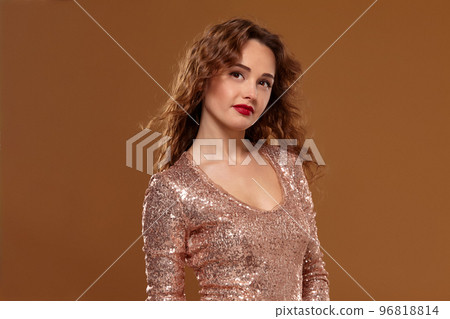 Portrait of young caucasian female in golden cocktail dress looking into the camera on brown background Portrait of young caucasian female in golden cocktail dress looking into the camera on brown background 96818814