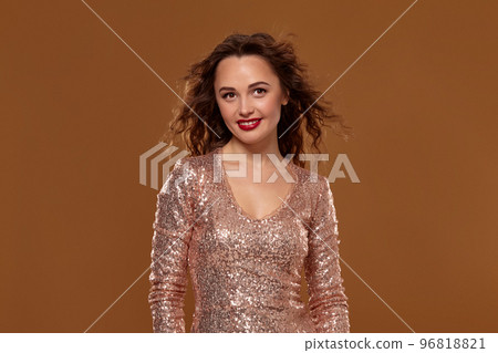 Portrait of young caucasian female in golden cocktail dress looking into the camera on brown background Portrait of young caucasian female in golden cocktail dress looking into the camera on brown background 96818821