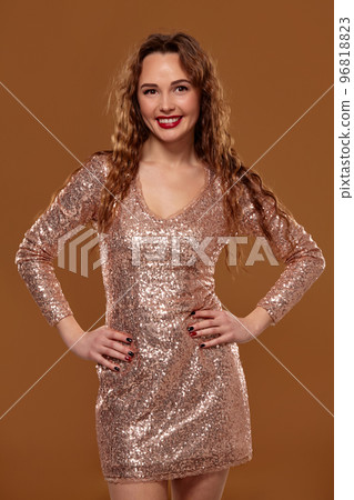 Portrait of young caucasian female in golden cocktail dress looking into the camera on brown background Portrait of young caucasian female in golden cocktail dress looking into the camera on brown background 96818823