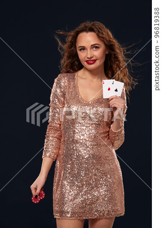 Brown-haired attractive caucasian young woman in golden cocktail dress at casino holds pair of aces and chips in her hands Brown-haired attractive caucasian young woman in golden cocktail dress at casino holds pair of aces and chips in her hands 96819388