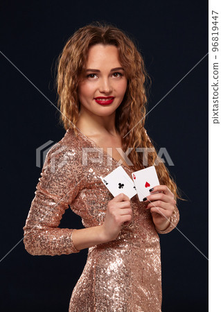 Pretty young brown-haired woman in golden cocktail dress holding pair of aces and smiling 96819447