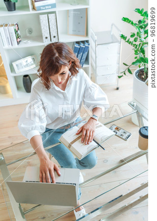 Woman in the office at the computer. She is dressed in a white shirt and blue jeans sitting at a table on which is a laptop and a notepad. Works in the office, bright workplace. Woman in the office at the computer. She is dressed in a white shirt and blue jeans sitting at a table on which is a laptop and a notepad. Works in the office, bright workplace. 96819699