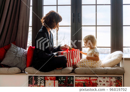 Mother and daughter sit on the windowsill and hold a gift between them. In a decorated Christmas room, mother in black pajamas, daughter in white dress. New Year's family holidays. Mother and daughter sit on the windowsill and hold a gift between them. In a decorated Christmas room, mother in black pajamas, daughter in white dress. New Year's family holidays. 96819702