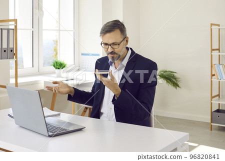 Mature businessman sitting at office desk and sending voice message on mobile phone 96820841