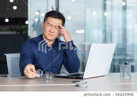 Asian businessman boss sick has severe headache, man in glasses sitting at desk using laptop at work inside office, migraine office worker investor overtired. 96820996