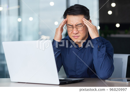 Asian businessman boss sick has severe headache, man in glasses sitting at desk using laptop at work inside office, migraine office worker investor overtired. 96820997